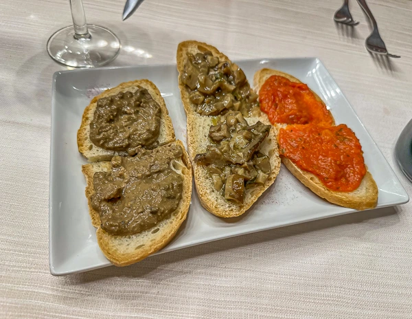 An image of a plate of crostini, featuring slices of toasted bread topped with different spreads. Three of the slices are topped with a greenish-brown mushroom and liver pâté, while the other two are covered with a bright red tomato sauce.