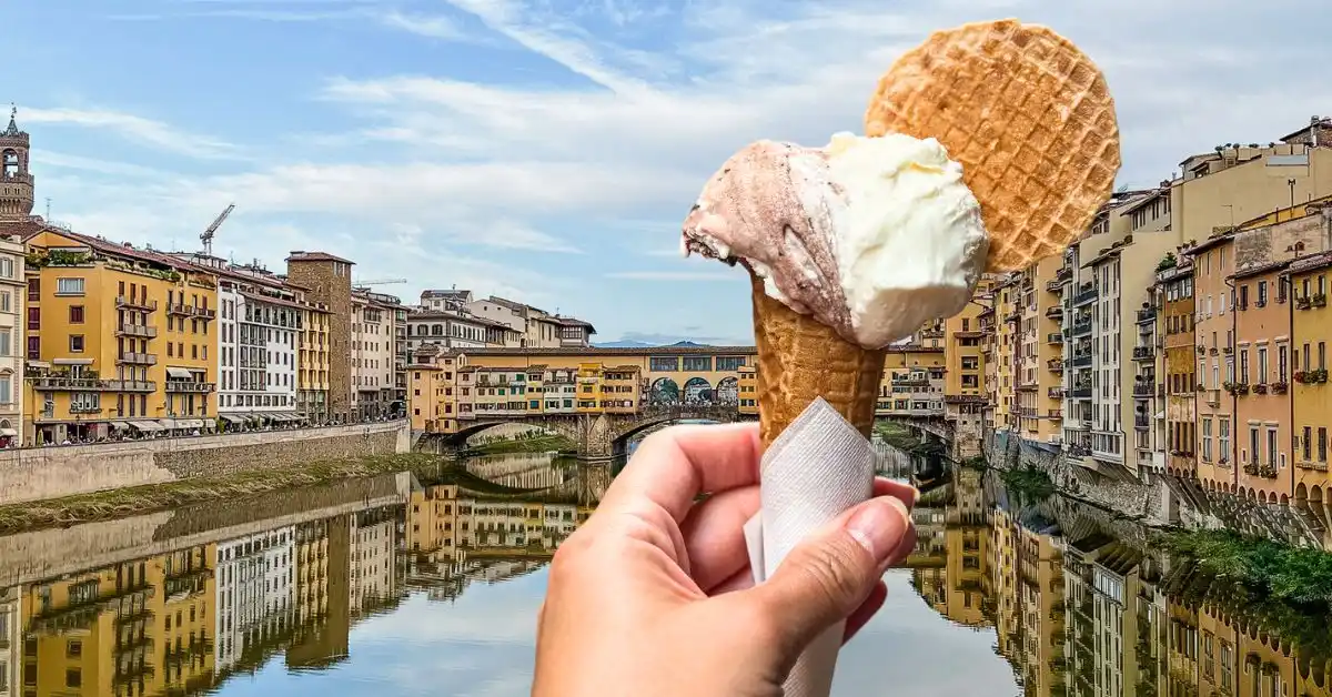 featured blog image of a hand holding a gelato cone with two scoops and a wafer, with the iconic Ponte Vecchio and reflective Arno River in the background, in Florence, Italy.