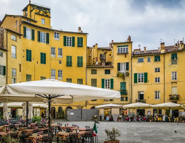 dining tables and umbrellas outside of a square surrounding by old yellow buildings in lucca italy