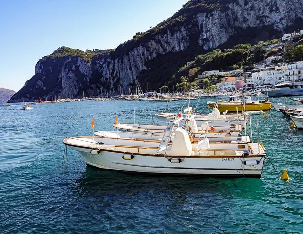 Small boats docked in the clear blue water off the coast of Capri, Italy, with rocky cliffs and hillside buildings in the background.