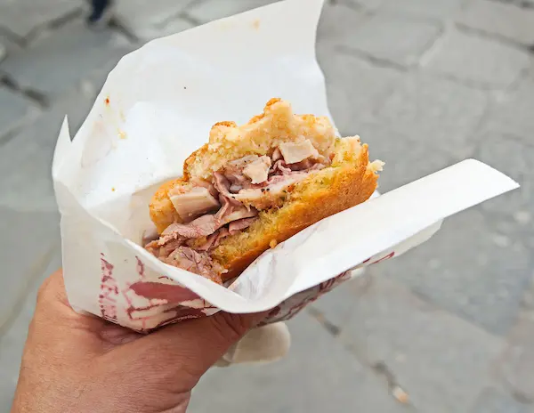 A close-up of a hand holding a trippa alla Fiorentina sandwich, served in a white paper wrapper. The sandwich is filled with tender pieces of tripe on Schiacciata Bread.