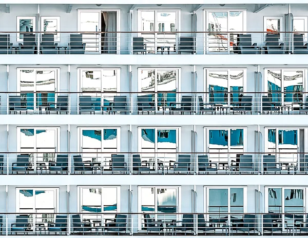 a detailed view of multiple balconies on a cruise ship, arranged in a repetitive, grid-like pattern. Each balcony is bordered by metal railings and includes outdoor chairs