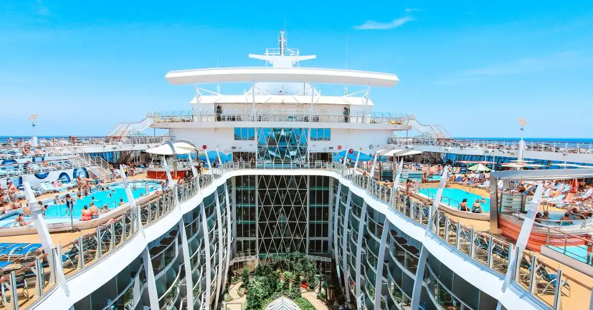 featured blog image without text | The image is a vibrant, clear day view of a cruise ship deck bustling with activity. It features a central atrium with a glass and white steel structure, surrounded by multiple levels of decks lined with loungers where guests are sunbathing and enjoying the pool area. The ship's radar and navigation equipment are visible atop the structure, set against a serene blue sky and calm sea. The services rendered on a cruise require cruise gratuities