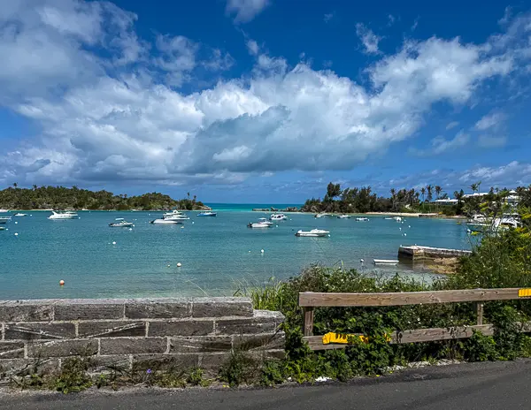 Harbor in Bermuda with turquoise water, small boats, stone wall, and shoreline scenery under a partly cloudy sky.
