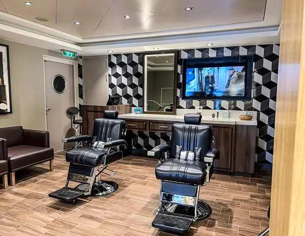 The image displays an interior view of a cruise ship's barber shop. There are two black leather barber chairs facing a large mirror, with a geometric black and white patterned wall providing a modern backdrop. The shop is equipped with a wooden floor, a waiting area with brown leather seats, and a flat-screen TV mounted above a wooden counter