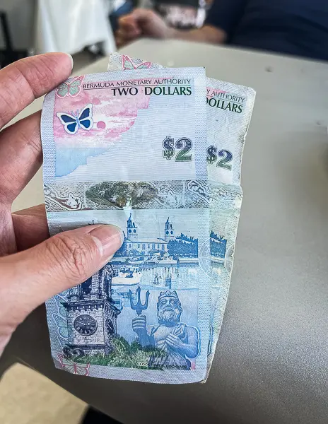 Two Bermuda two-dollar bills held in hand, showing Bermuda currency with colorful island artwork and the Bermuda Monetary Authority name.