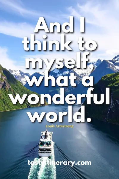 Cruise ship sailing through a narrow fjord surrounded by steep green mountains and snow-capped peaks under a blue sky. Text on the image reads: “And I think to myself, what a wonderful world. – Louis Armstrong.”