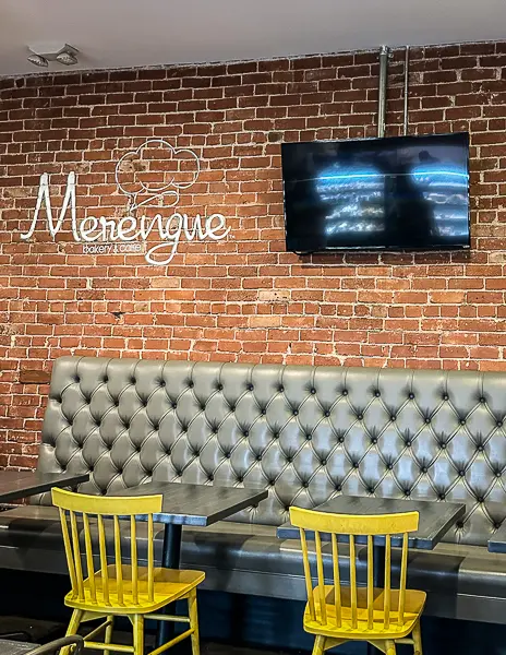 Indoor seating area at Merengue Bakery & Cafe with tufted booth, yellow chairs, and brick wall featuring the Merengue logo.
