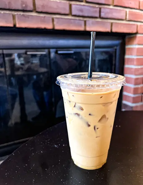 An iced coconut brevi from Jameson Brown Coffee Roasters, served in a clear to-go cup with a straw, placed on a table near a brick fireplace.