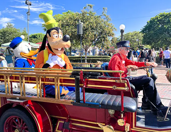 Goofy and Donald Duck ride on a vintage fire truck down Main Street, U.S.A., at Disneyland, with a driver dressed in red leading the way.