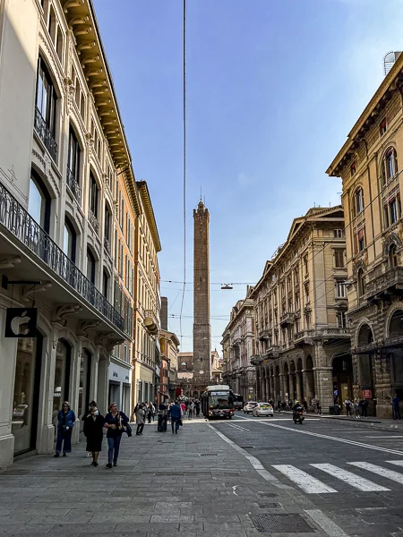 A distant view of Torre degli Asinelli in Bologna, Italy, seen from a wide street lined with elegant historic buildings.