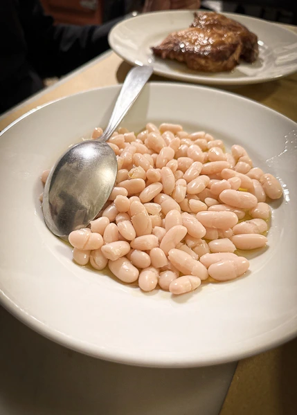 a bowl of Fagioli all'olio, featuring creamy and tender white beans served in a white bowl with a spoon, suggesting a simple yet delicious Italian dish. In the background, there's a glimpse of another plate, possibly with meat