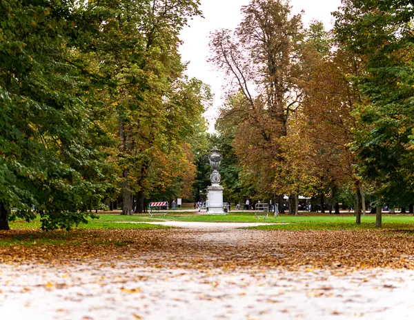 Leaf-covered path through Parco Ducale in Parma, Italy, with autumn trees lining the walkway and a statue visible in the distance