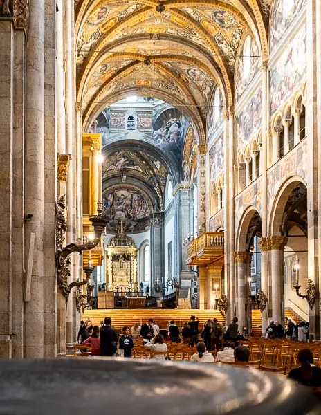 View down the central nave of Parma Cathedral with golden vaulted ceilings, painted walls, and people seated below