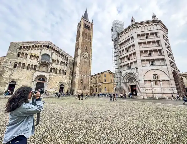 Tourist taking a photo in Piazza Duomo in Parma, Italy, with the Parma Cathedral, bell tower, and Baptistery in view on a cloudy day