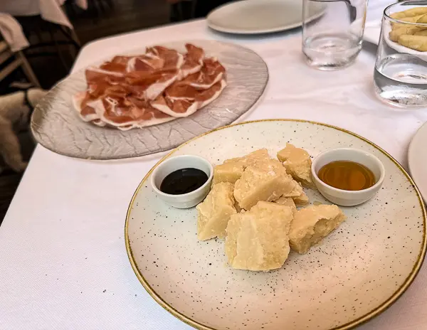 Plate of Parmigiano Reggiano chunks served with balsamic vinegar and honey at La Forchetta restaurant in Parma, with Prosciutto di Parma in the background
