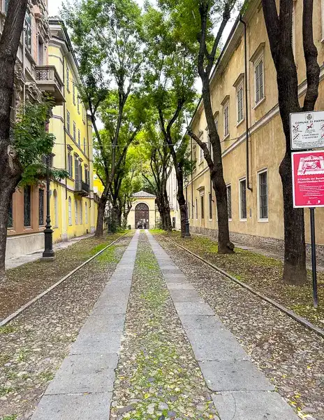 Cobblestone and stone pathway lined with trees between colorful buildings in Parma, Italy