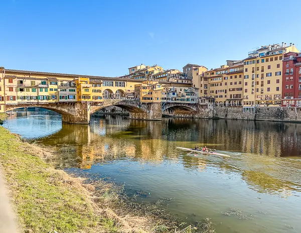 View of the Ponte Vecchio spanning the Arno River in Florence, Italy, with rowers gliding through the water below.