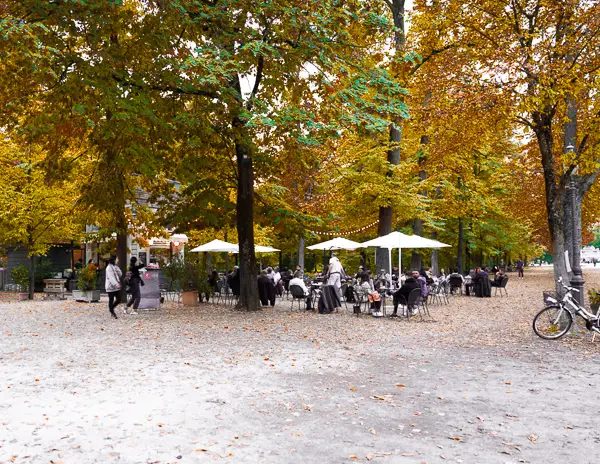 People sitting at outdoor tables under umbrellas at daMAT Chiosco café in Parco Ducale, Parma, surrounded by autumn trees