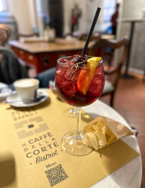 A vibrant blackberry spritz cocktail garnished with an orange slice and berries, served with a side of chips at Il Caffè Della Corte Bistrot in Bologna.