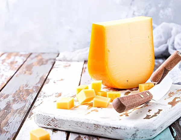 A block of golden yellow cheese on a rustic white cutting board, surrounded by small cheese cubes and a cheese knife with a wooden handle. The background features a light gray and wooden surface.