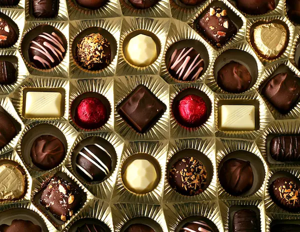A close-up view of an assorted box of chocolates, featuring a variety of shapes, textures, and toppings, displayed in gold-lined compartments.