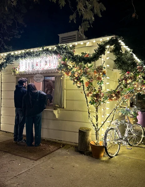 Hot cocoa stand at 123 Farm, adorned with Christmas garlands and lights, as two people place their order on a cold winter night.