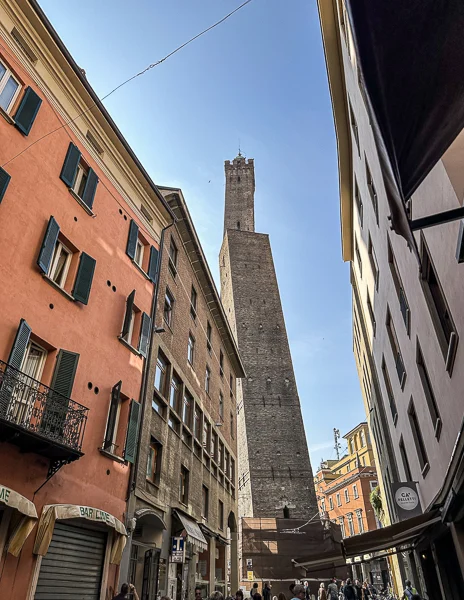 A street-level view of Torre degli Asinelli and Torre Garisenda in Bologna, Italy, framed by colorful buildings and bustling streets.