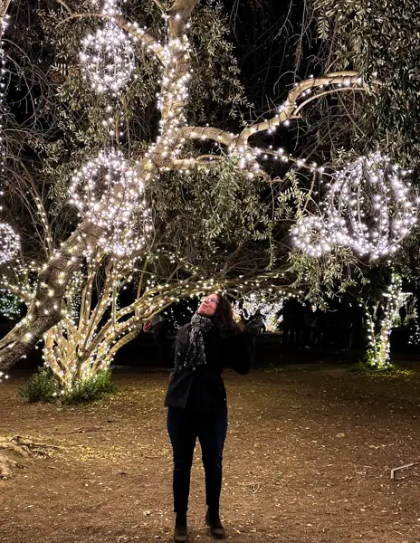A woman standing beneath a tree decorated with glowing spherical lights at 123 Farm during Christmastime, looking up and enjoying the festive holiday display.