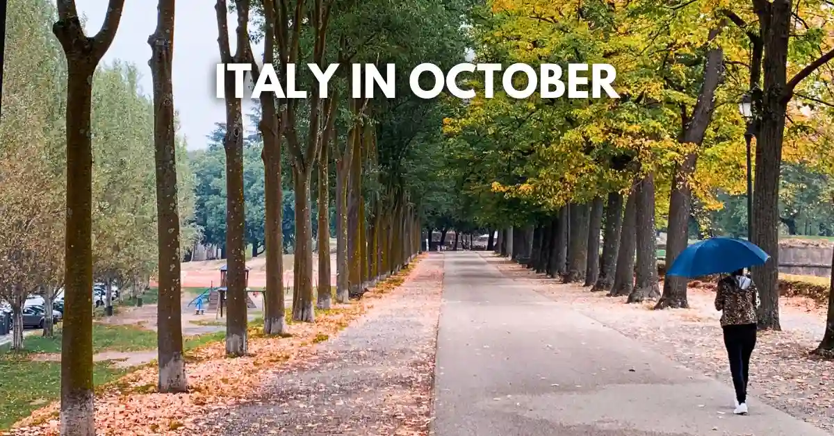 featured blog image: A tree-lined path in Italy during early autumn, with fallen leaves scattered along the ground. A person walks away holding a bright blue umbrella under overcast skies. Text on the image reads: “Italy in October.”