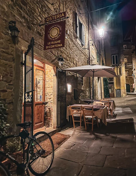 The charming exterior of Osteria Le Rughe di Cortona, Italy, at night, with a softly lit entrance, outdoor seating, and cobblestone streets creating a warm, inviting atmosphere.