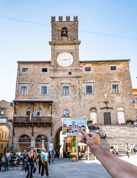 A hand holding a colorful postcard of Cortona in front of the Palazzo Comunale, the iconic town hall with its clock tower and stone steps, in Piazza della Repubblica.