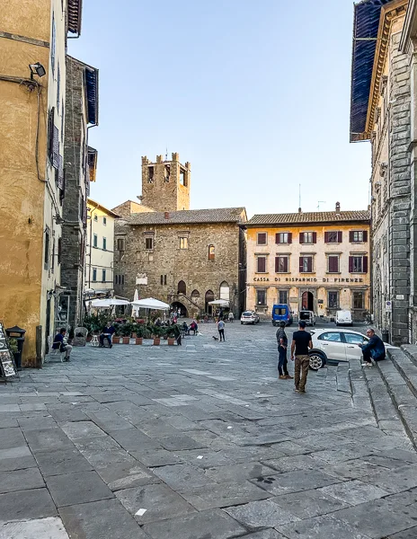 A view of Piazza Signorelli in Cortona, Italy, with the Signorelli Theatre, stone buildings, and outdoor seating under a clear blue sky, capturing the charm of the square.