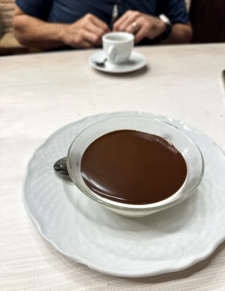 A small glass dish of budino al cioccolato, a rich panna cotta with chocolate dessert served at Trattoria Dardano in Cortona, Italy, with a cup of espresso in the background.