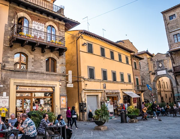 A lively scene at Piazza della Repubblica in Cortona, Italy, featuring outdoor cafés with people dining, historic buildings, and colorful storefronts under a sunny sky.