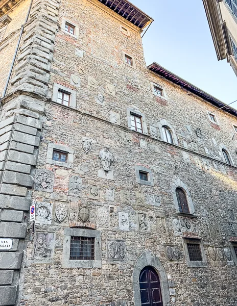 A historic stone building in Piazza Signorelli, Cortona, Italy, adorned with an array of carved coats of arms and decorative stonework, showcasing its rich history.