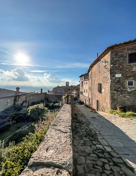 A scenic view of Cortona's cobblestone streets and stone buildings bathed in sunlight, with rolling Tuscan hills in the background.