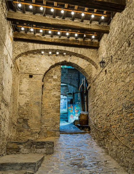 A charming stone archway illuminated by string lights, leading to a narrow cobblestone alley in Cortona, Italy, with colorful artwork visible in the distance.