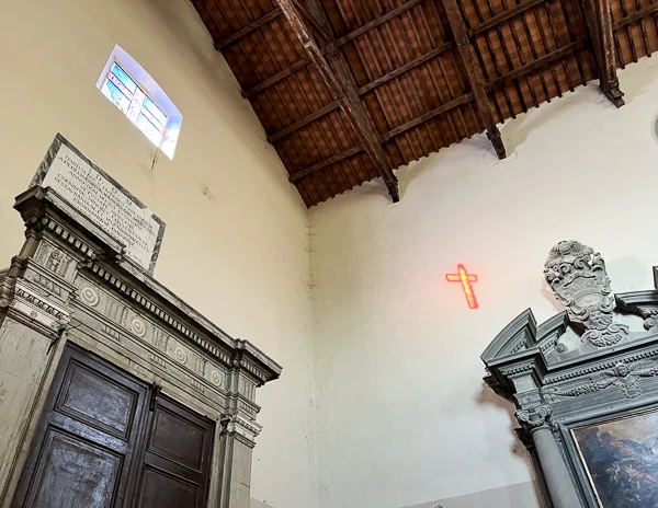 The interior of the Convento di San Francesco in Cortona, Italy, showing a stained-glass window casting a glowing red cross on the wall, creating a serene and reflective atmosphere.