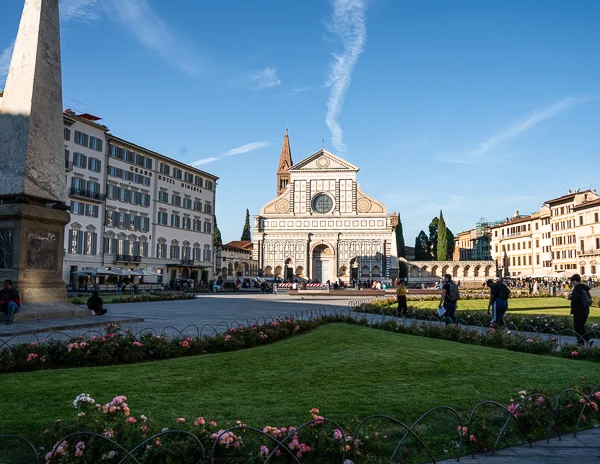 The Basilica of Santa Maria Novella in Florence, Italy, featuring its stunning Renaissance and Gothic-style facade, with a vibrant green lawn and colorful flowers in the foreground under a bright blue sky.