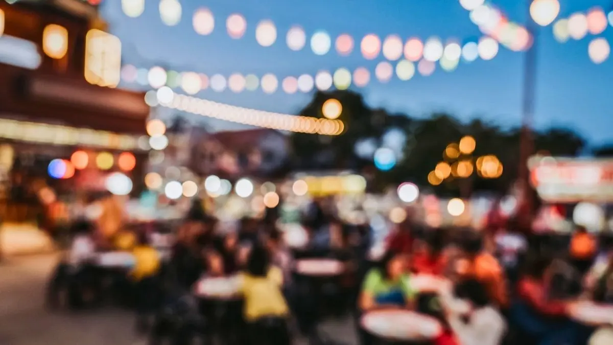 featured image: Blurred outdoor dining scene at dusk, with people seated at restaurant tables beneath colorful string lights, used as the featured image for a collection of food and travel quotes.
