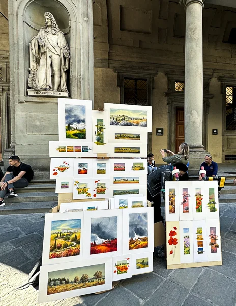 A display of vibrant paintings and artwork by local artists set up outside the Uffizi Gallery in Florence, Italy, with the statue of Francesco Redi in the background.