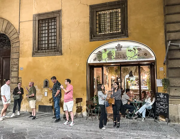 The exterior of Babae in Florence, Italy, featuring its iconic wine window (Buchette del Vino) to the left of the entrance, with people gathered outside enjoying drinks and the cozy ambiance of the cafe.
