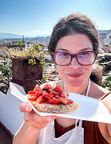 A woman (kathy) holding a plate of freshly made bruschetta with tomatoes during a cooking class in Florence, with a rooftop view of the city in the background.