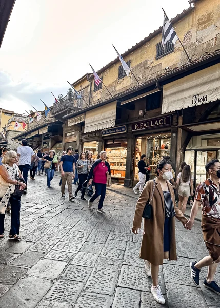 Crowds walking across the historic Ponte Vecchio bridge in Florence, Italy, lined with shops and flags hanging above.
