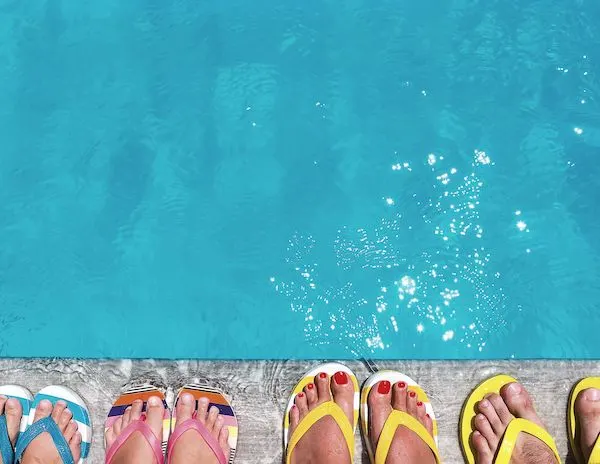 4 pairs of feet wearing flip flops of different colors next to a pool