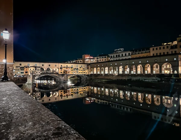 A detailed nighttime shot of Ponte Vecchio in Florence, with its arches and windows perfectly mirrored on the Arno River, surrounded by the city’s glowing lights.