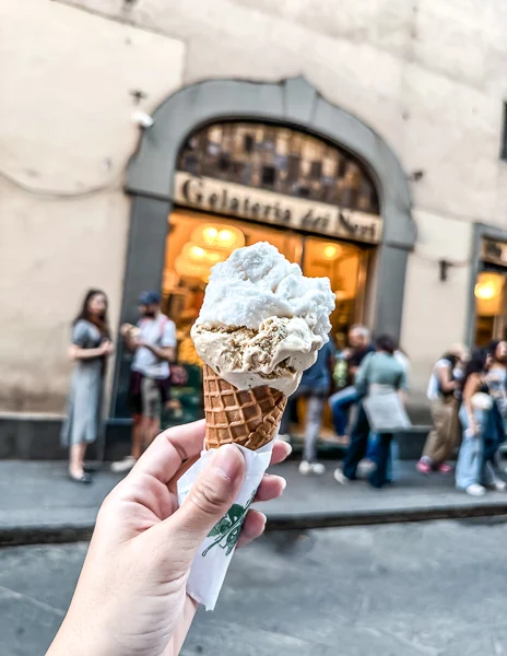 A hand holding a waffle cone with two scoops of gelato in front of Gelateria dei Neri in Florence, Italy, with the shop’s entrance and a small crowd in the background.