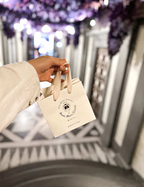 A shopper holding a small bag from the Farmaceutica di Santa Maria Novella in Florence, showcasing its iconic logo. The elegant interior design is visible in the blurred background.
