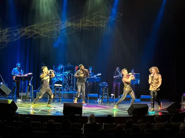 4 men singing on stage with band in backround